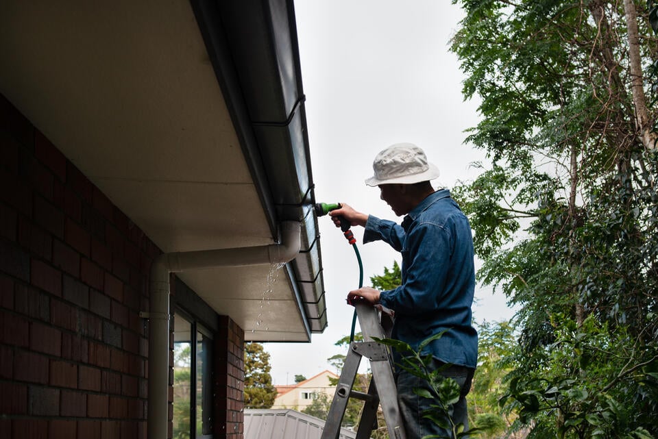 diy gutter cleaning-man standing on a ladder and using hose to clean gutter system on house