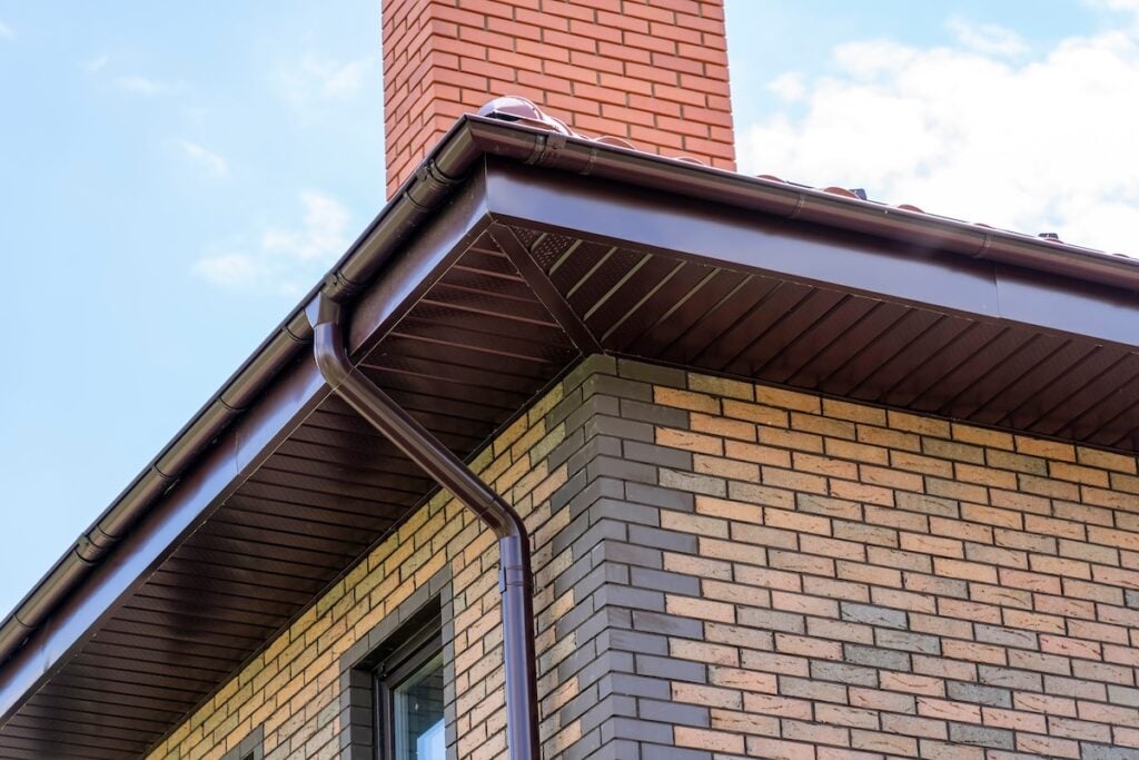 close-up view of house with brown roof and filing of roof overhangs with soffits and brick stone chimney pipe
