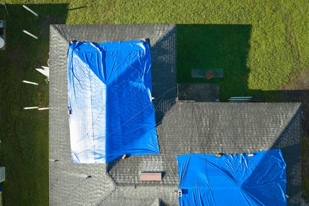 how to put a tarp on a roof without nails Top view of leaking house roof covered with protective tarp sheets against rain water leaks until replacement of asphalt shingles. Damage of building rooftop as aftermath