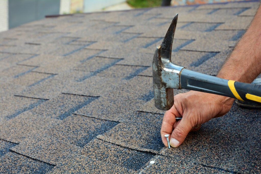 where to nail shingles Worker hands installing bitumen roof shingles. Worker Hammer in Nails on the Roof. Roofer is hammering a Nail in the Roof Shingles. Unfinished roof.