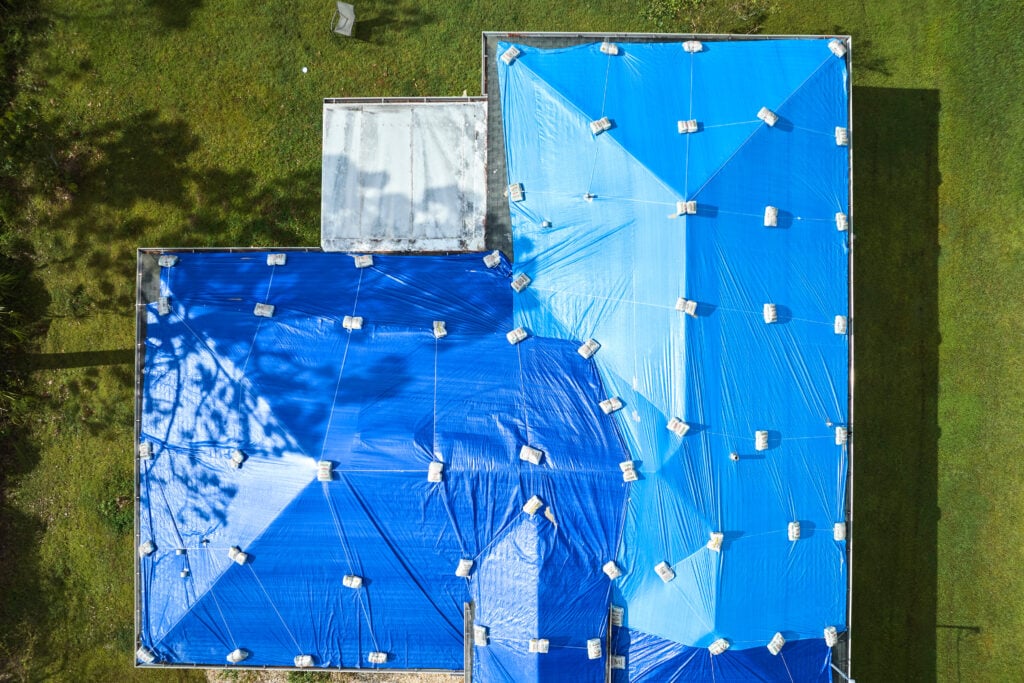 how to put a tarp on a roof without nails Top view of leaking house roof covered with protective tarp sheets against rain water leaks until replacement of asphalt shingles. Damage of building rooftop as aftermath