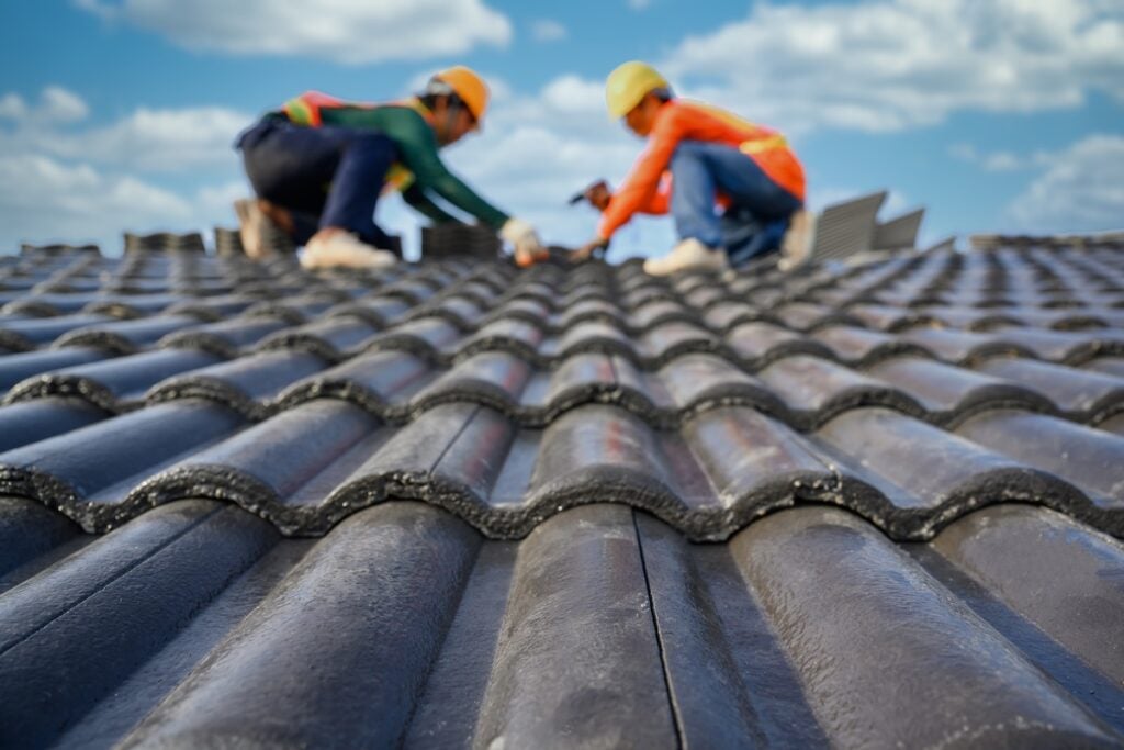 questions to ask a roofer Select the focus on roof tiles. Blurred background of a roofer working on the roof of a house Use a drill to drill the screws to fix the cement tiles.