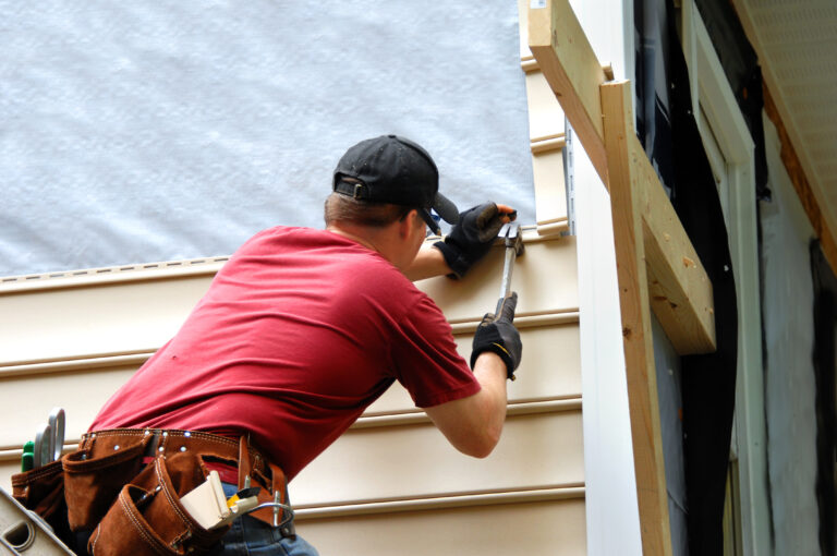 Young homeowner installs siding to his home. He is holding a hammer and wearing a tool belt.