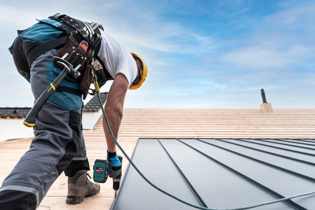 questions to ask a roofer A roofer with a safety harness and tool belt is working with a electric screwdriver on the roof. He is anchoring the metal roofing with a screws.