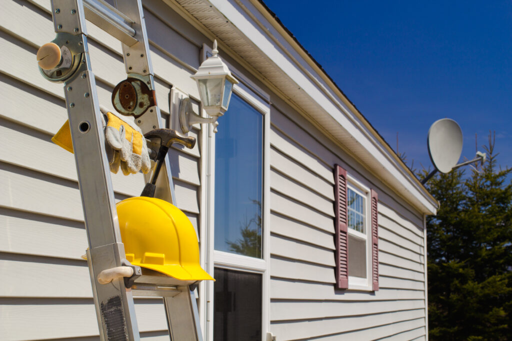 siding repair cost Yellow hard hat, work gloves and hammer on ladder with house and wireless dish background.