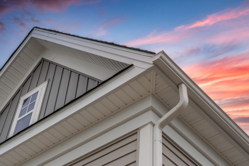 White frame gutter guard system, with gray horizontal and vertical vinyl siding fascia, drip edge, soffit, on a pitched roof attic at a luxury American single family home dramatic sunset sky