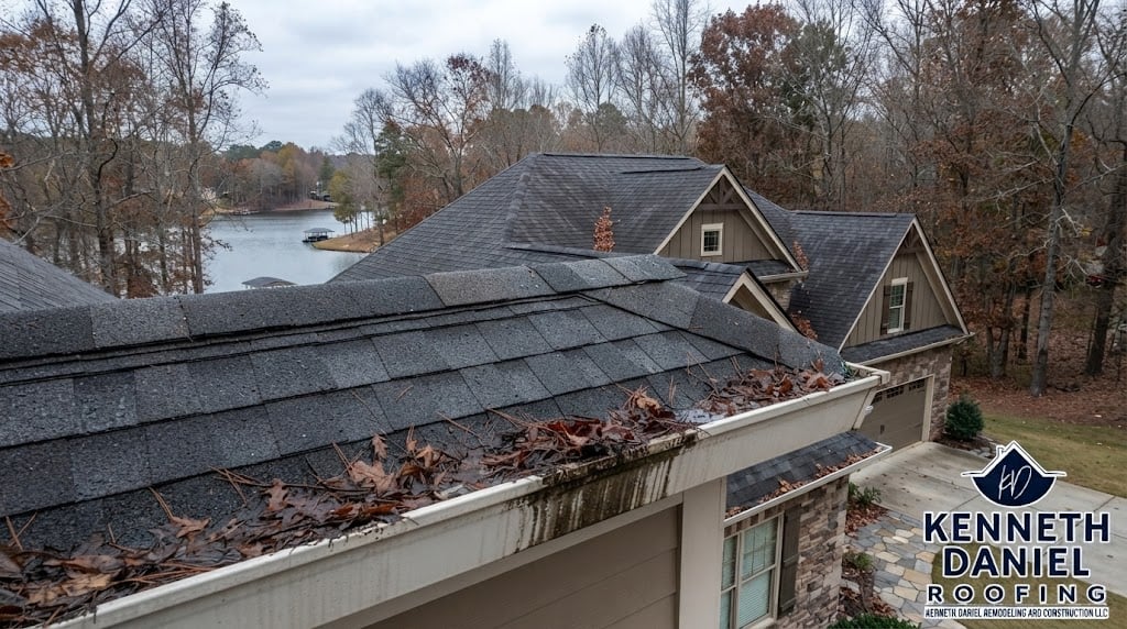 High-angle photo of a residential lake house roof in late fall showing heavy accumulation of dry leaves and pine needles in the white gutters, indicating the need for cleaning.